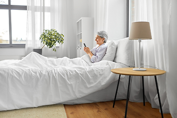 Image showing happy senior woman using smartphone in bed at home