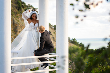 Image showing The newlyweds are walking in a beautiful picturesque gazebo, the girl is holding a veil with her hand, the guy is sitting on the railing