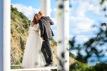 Image showing Happy newlyweds kiss in a beautiful gazebo with columns against the backdrop of foliage and sky