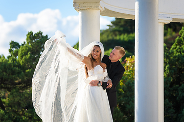 Image showing Happy newlyweds hugging on a walk, the girl raised her veil with her hand and smiles