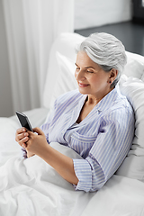 Image showing happy senior woman using smartphone in bed at home