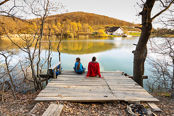 Image showing A girl and a girl are sitting on a wooden bridge near a mountain lake, a view from the back of people