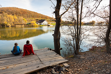 Image showing A girl and a girl are sitting on a wooden bridge by the lake and looking at a beautiful autumn landscape