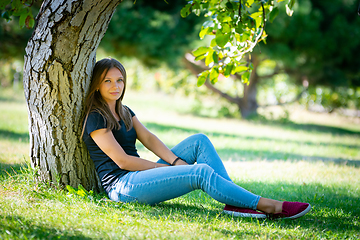 Image showing A girl sits under a tree in a sunny park and looks into the frame