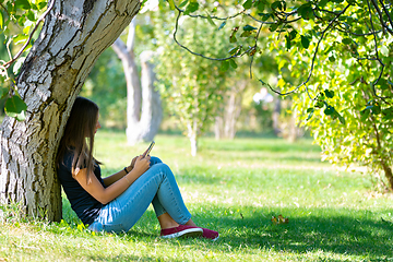 Image showing A girl sits under a tree in a sunny park and looks at the phone screen