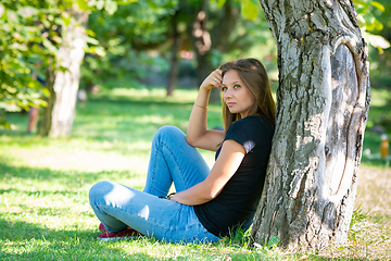 Image showing A girl sits under a tree in a sunny park and looks into the distance in thought