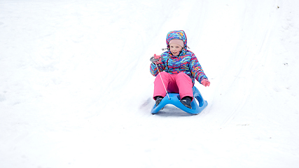 Image showing Cheerful girl riding a sled downhill on a snow covered sledge trail in a white sunny winter mountain landscape