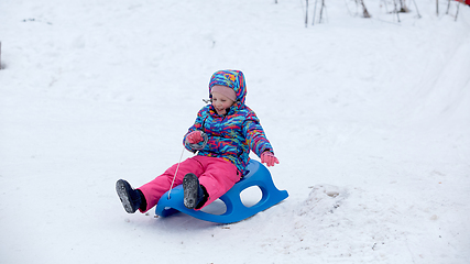 Image showing Cheerful girl riding a sled downhill on a snow covered sledge trail in a white sunny winter mountain landscape