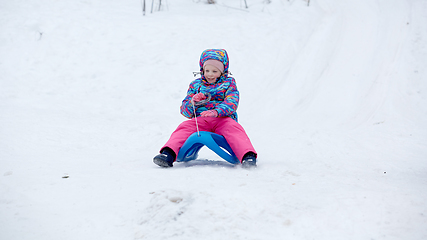 Image showing Cheerful girl riding a sled downhill on a snow covered sledge trail in a white sunny winter mountain landscape