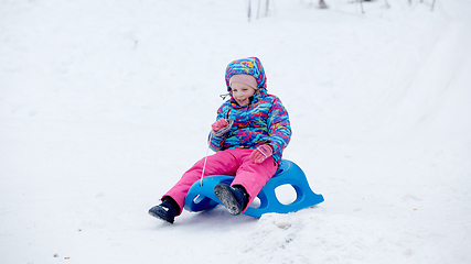 Image showing Cheerful girl riding a sled downhill on a snow covered sledge trail in a white sunny winter mountain landscape