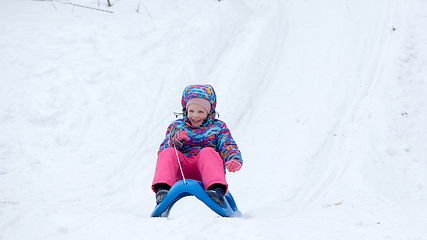 Image showing Cheerful girl riding a sled downhill on a snow covered sledge trail in a white sunny winter mountain landscape