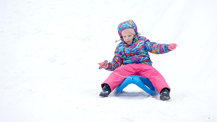 Image showing Cheerful girl riding a sled downhill on a snow covered sledge trail in a white sunny winter mountain landscape