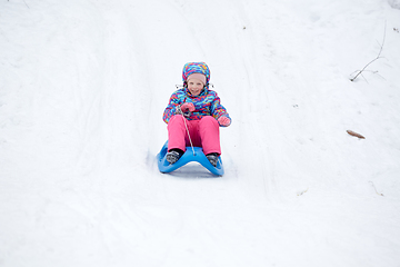 Image showing Cheerful girl riding a sled downhill on a snow covered sledge trail in a white sunny winter mountain landscape