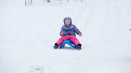 Image showing Cheerful girl riding a sled downhill on a snow covered sledge trail in a white sunny winter mountain landscape