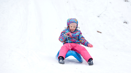 Image showing Cheerful girl riding a sled downhill on a snow covered sledge trail in a white sunny winter mountain landscape