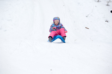 Image showing Cheerful girl riding a sled downhill on a snow covered sledge trail in a white sunny winter mountain landscape