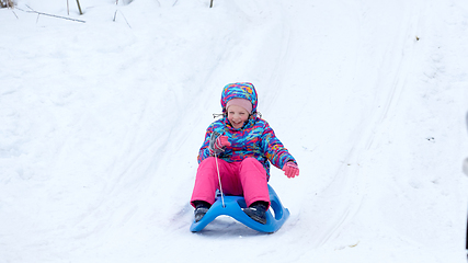 Image showing Cheerful girl riding a sled downhill on a snow covered sledge trail in a white sunny winter mountain landscape