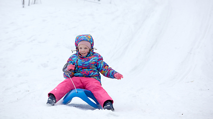 Image showing Cheerful girl riding a sled downhill on a snow covered sledge trail in a white sunny winter mountain landscape