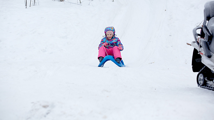 Image showing Cheerful girl riding a sled downhill on a snow covered sledge trail in a white sunny winter mountain landscape