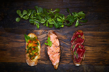 Image showing Assorted bruschetta with roast beef, vegetables and lightly salted salmon with greens leaves on wooden background.