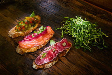 Image showing Assorted bruschetta with roast beef, vegetables and lightly salted salmon with greens leaves on wooden background.