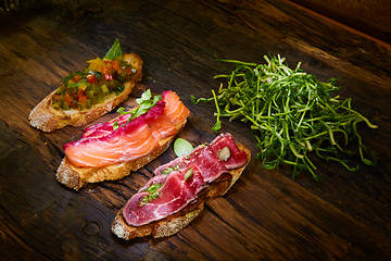 Image showing Assorted bruschetta with roast beef, vegetables and lightly salted salmon with greens leaves on wooden background.