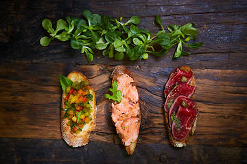 Image showing Assorted bruschetta with roast beef, vegetables and lightly salted salmon with greens leaves on wooden background.