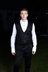 Image showing Male fashion, beauty concept. Portrait of young man with short wet hair wearing black suit, posing on street. Classic style.