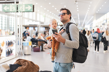 Image showing Father traveling with child, holding his infant baby boy at airport terminal, checking flight schedule, waiting to board a plane. Travel with kids concept.