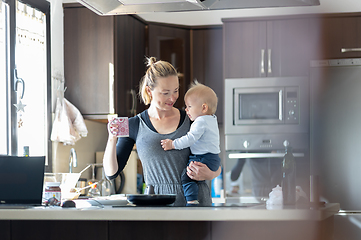 Image showing Happy mother holding her little infant baby boy while drinking morning coffee and making pancakes for breakfast in domestic kitchen. Family lifestyle, domestic life concept.