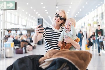 Image showing Mother taking selfie with mobile phone, while traveling with child, holding his infant baby boy at airport, waiting to board a plane. Travel with kids concept.