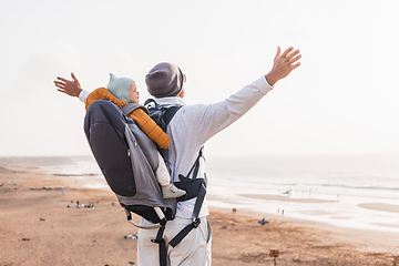 Image showing Young father rising hands to the sky while enjoying pure nature carrying his infant baby boy son in backpack on windy sandy beach. Family travel concept.