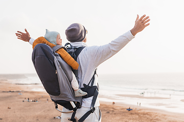 Image showing Young father rising hands to the sky while enjoying pure nature carrying his infant baby boy son in backpack on windy sandy beach. Family travel concept.
