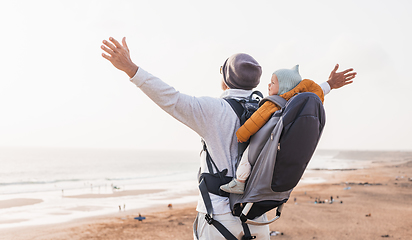 Image showing Young father rising hands to the sky while enjoying pure nature carrying his infant baby boy son in backpack on windy sandy beach. Family travel concept.