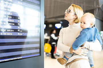 Image showing Mother traveling with child, holding his infant baby boy at airport terminal, checking flight schedule, waiting to board a plane. Travel with kids concept.