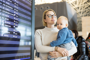 Image showing Mother traveling with child, holding his infant baby boy at airport terminal, checking flight schedule, waiting to board a plane. Travel with kids concept.