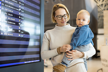 Image showing Mother traveling with child, holding his infant baby boy at airport terminal, checking flight schedule, waiting to board a plane. Travel with kids concept.