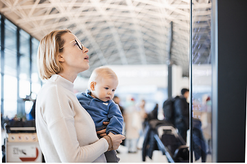 Image showing Mother traveling with child, holding his infant baby boy at airport terminal, checking flight schedule, waiting to board a plane. Travel with kids concept.