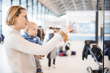 Image showing Mother traveling with child, holding his infant baby boy at airport terminal, checking flight schedule, waiting to board a plane. Travel with kids concept.