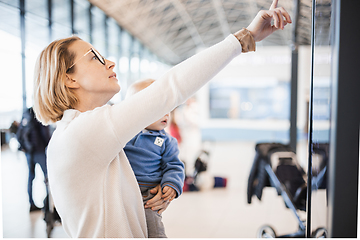 Image showing Mother traveling with child, holding his infant baby boy at airport terminal, checking flight schedule, waiting to board a plane. Travel with kids concept.