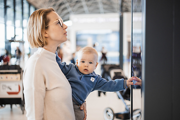 Image showing Mother traveling with child, holding his infant baby boy at airport terminal, checking flight schedule, waiting to board a plane. Travel with kids concept.