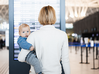 Image showing Mother traveling with child, holding his infant baby boy at airport terminal, checking flight schedule, waiting to board a plane. Travel with kids concept.