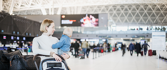 Image showing Motherat travelling with his infant baby boy child, walking, pushing baby stroller and luggage cart at airport terminal station. Travel with child concept.