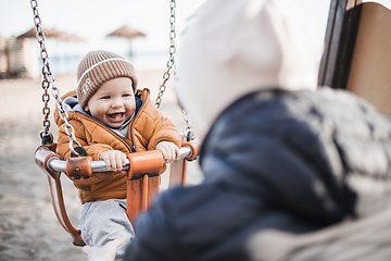 Image showing Mother pushing her cheerful infant baby boy child on a swing on sandy beach playground outdoors on nice sunny cold winter day in Malaga, Spain.