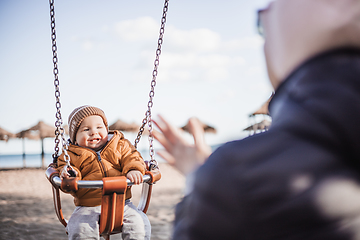 Image showing Mother pushing her cheerful infant baby boy child on a swing on sandy beach playground outdoors on nice sunny cold winter day in Malaga, Spain.