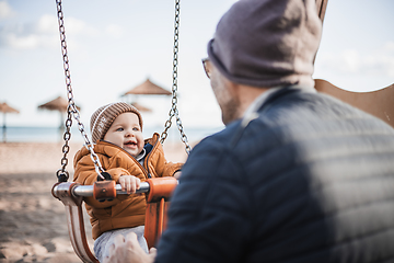 Image showing Father pushing hir cheerful infant baby boy child on a swing on sandy beach playground outdoors on nice sunny cold winter day in Malaga, Spain.