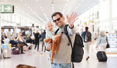 Image showing Father traveling with child, holding his infant baby boy at airport terminal waiting to board a plane waving goodby. Travel with kids concept.