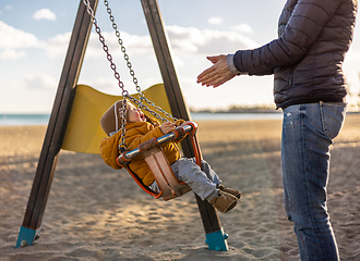 Image showing Mother pushing her infant baby boy child on a swing on sandy beach playground outdoors on nice sunny cold winter day in Malaga, Spain.