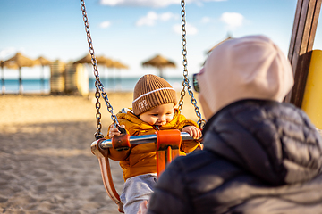 Image showing Mother pushing her infant baby boy child on a swing on sandy beach playground outdoors on nice sunny cold winter day in Malaga, Spain.