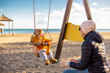 Image showing Mother pushing her infant baby boy child on a swing on sandy beach playground outdoors on nice sunny cold winter day in Malaga, Spain.